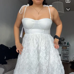 Vit klänning  - Säljer en söt vit klänning med axelband och blommigt mönster på kjolen. Klänningen har en tight passform upptill med en hjärtformad urringning och en luftig kjol. Perfekt för studenten! 