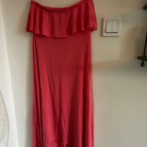 NY- axelbandslös klänning från Gina Tricot - Klänningen är aldrig använd. Kolla på anta bilden för att se hur den ser ut ner till. Snygg rosa axelbandslös klänning från Gina Tricot med volangdetalj upptill. Klänningen har en luftig passform och är tillverkad i ett mjukt material. FINNS FLER BILDER