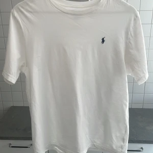 Vit t-shirt från Ralph Lauren - Säljer en klassisk vit t-shirt från Ralph Lauren med det ikoniska broderade logotypen på bröstet. T-shirten har en normal passform och är tillverkad i mjuk bomull. Perfekt för en stilren och avslappnad look. Sitter som en M