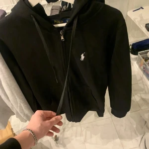 Svart hoodie från Polo Ralph Lauren - Säljer en stilren svart hoodie från Polo Ralph Lauren med dragkedja och det klassiska logotypbroderiet på bröstet. Perfekt för en avslappnad look. Den har en justerbar huva och långa ärmar.