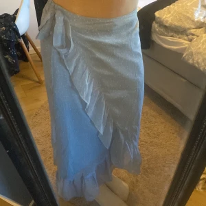 Ljusblå omlottkjol med volang - Säljer en söt ljusblå omlottkjol med volangdetaljer och knytning i midjan. Kjolen har ett diskret prickigt mönster och är perfekt för vår och sommar. Den är i en längre modell och ger en romantisk känsla. Passar utmärkt till både vardag och festliga tillfällen.