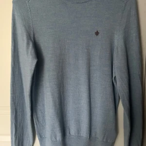 Ljusblå tröja från Morris - Säljer en stilren ljusblå tröja från Morris i 100% merinoull. Tröjan har en liten broderad detalj på bröstet och ribbade kanter vid ärmslut och nederkant. Perfekt för en elegant och avslappnad look. Sitter ganska tajt för en S så passar också XS