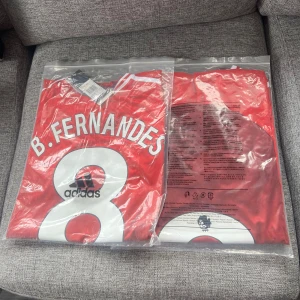 Röd fotbollströja från Adidas - Säljer en röd fotbollströja från Adidas med B. Fernandes och nummer 8 på ryggen. Tröjan har vita detaljer och är kortärmad. Perfekt för fotbollsfans!