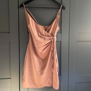 Champagnefärgad omlottklänning - Champagnefärgad klänning från BikBok i storlek XS. Omlott-design med knytning i sidan och smala axelband. Dragkedja baktill.