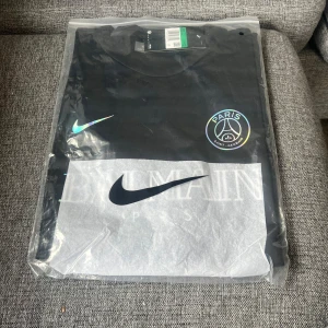 Svart PSG fotbollströja från Nike - Säljer en svart PSG fotbollströja från Nike med klubbens emblem på bröstet och ett turkost Nike-märke. Tröjan är kortärmad och perfekt för alla fotbollsfans. Levereras i originalförpackning.