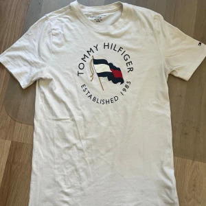 Vit t-shirt från Tommy Hilfiger - Säljer en stilren vit t-shirt från Tommy Hilfiger med deras ikoniska logga och texten 'Established 1985' tryckt på framsidan. T-shirten har korta ärmar och är tillverkad i mjuk bomull. Perfekt för en avslappnad look! Nästan helt ny och storlek XL hos barn.