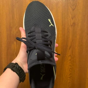 Svarta sneakers från Puma - Snygga svarta sneakers från Puma med vit sula och märkets logga i gult på sidan och tån. Skorna har snörning och en modern design som passar perfekt för träning eller vardagsbruk.