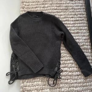 Mörkgrå stickad tröja med snörning - Säljer en snygg mörkgrå stickad tröja med cool snörning på sidorna. Tröjan har långa ärmar och en rund halsringning. Perfekt för kyliga dagar!