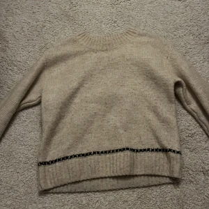 Beige stickad tröja med mönster - Säljer en mysig beige stickad tröja med ett subtilt mönster i svart längs ärmarna och nederkanten. Perfekt för kyliga dagar.