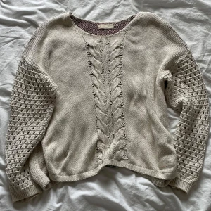 Beige stickad tröja från Odd Molly - Säljer en beige stickad tröja från Odd Molly med flätstickat mönster framtill och ett färgglatt broderat mönster på ryggen i rosa, svart och turkos. Tröjan har långa ärmar med hålmönster och rund halsringning. Perfekt för att ge din outfit en unik touch! 