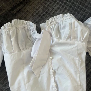 Vit bandeu blus med puffärm - Vit blus med puffärmar och volanger över bysten. Blusen har en knäppning framtill med pärlknappar och en dekorativ knytning. Strl: 42/L Prettylittlething (PLT) (aldrig använt) 