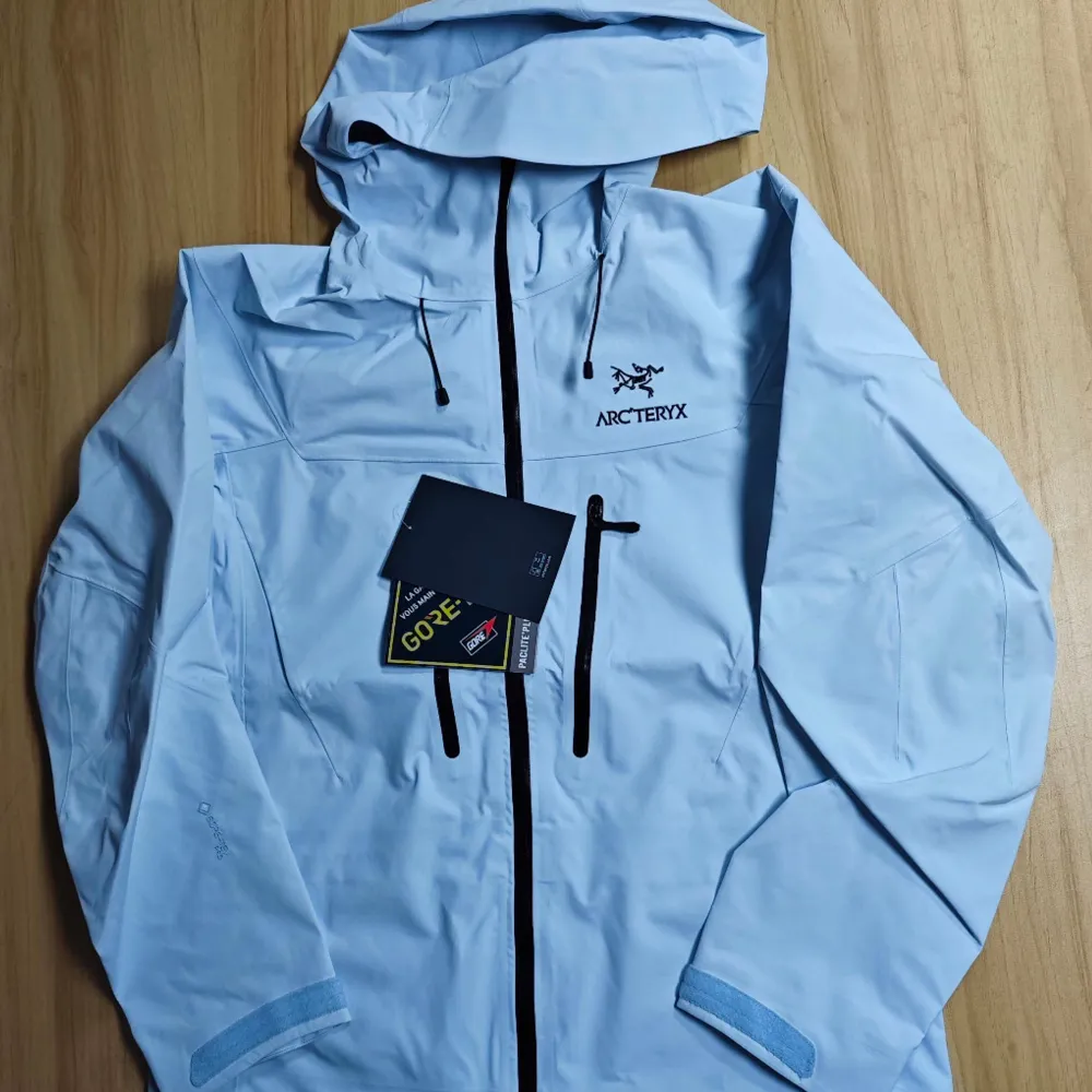 Säljer en ljusblå vindjacka från Arc'teryx med huva och dragkedja. Jackan har flera praktiska fickor med dragkedjor och är perfekt för utomhusaktiviteter. Den är tillverkad i ett slitstarkt material och har en stilren design.. Takit.