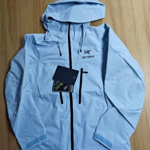 Ljusblå vindjacka från Arc'teryx - Säljer en ljusblå vindjacka från Arc'teryx med huva och dragkedja. Jackan har flera praktiska fickor med dragkedjor och är perfekt för utomhusaktiviteter. Den är tillverkad i ett slitstarkt material och har en stilren design.