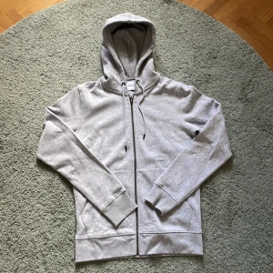 Grå hoodie från Jack & Jones - Säljer en stilren grå hoodie från Jack & Jones. Den har en dragkedja framtill och praktiska fickor. Perfekt för en avslappnad look och i arbetsmiljö. Smälter in bra! Nypris: 750 kr