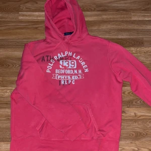 Röd hoodie från Polo Ralph Lauren XL - Snygg röda hoodie från Polo Ralph Lauren i storlek XL. Tröjan har vit tryckt text och siffror framtill, klassisk huva med snörning och en stor magficka. Perfekt för dig som gillar sportig och avslappnad stil.