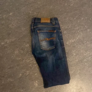 Nudie Jeans blå jeans W27 L32 - Snygga blå jeans från Nudie Jeans med klassisk femficksdesign och orangea kontrastsömmar. Jeansen har en rak passform och är tillverkade i slitstarkt denim. Bakfickan har Nudie Jeans ikoniska broderi. Perfekta för dig som gillar stilrena och tidlösa jeans.