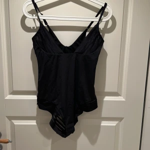 Svart body  - Snygg svart body med smala axelband och v-ringning både fram och bak. Bodyn har genomskinliga meshdetaljer vid höfterna som ger en cool look. Perfekt att styla med jeans eller kjol för en trendig outfit.