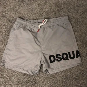 Gråa DSQUARED2 badshorts med tryck - Snygga grå badshorts från DSQUARED2 med svart logotyp-tryck runt benen. Shortsen har elastisk midja med vit snörning och röda detaljer samt innerbyxa i mesh. Perfekta för stranden eller poolen, riktigt clean look. OBS:sitter som xs, ganska korta