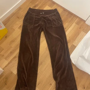 Bruna velourbyxor från Juicy Couture - Mysiga bruna mjukisbyxor från Juicy Couture i velour med broderad JC-logga och silvriga knappar på bakfickorna. Byxorna har normal passform, resår i midjan och raka ben. Perfekta för en chill och trendig look.