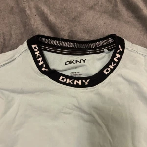Ljusblå DKNY t-shirt med logga - Snygg ljusblå t-shirt från DKNY med svart ribbad krage där DKNY-loggan är invävd i vitt. T-shirten har korta ärmar och tryckt logga på båda axlarna. Tillverkad i mjuk bomull, perfekt för en clean och stilren look.