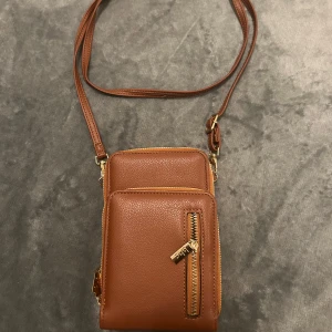 Brun axelväska i skinn med dragkedja - Snygg brun axelväska från BHC i slätt skinn med guldfärgade detaljer. Väskan har två separata fack med dragkedjor, ett ytterfack med dragkedja och flera kortfack inuti. Perfekt storlek för mobilen och det viktigaste.