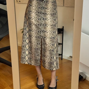 Beige långkjol med ormmönster - Säljer en beige och svart långkjol med snyggt ormmönster. Kjolen har en lätt glansig yta och sitter högt i midjan med en rak men något utsvängd passform. Perfekt för dig som vill sticka ut med en trendig look.