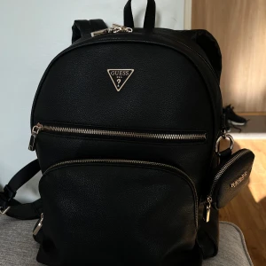 Svart Guess ryggsäck - Svart Guess ryggsäck med justerbara axelremmar, ett huvudfack med dragkedja, en framficka med dragkedja och en liten avtagbar liten väska. Ryggsäcken har en Guess-logotyp på framsidan och ett mönstrat foder med Guess-logotyper. Mycket rymlig och trendig. I mycket fint skick då varsam använd. Litet märke på den lilla väskan, se bild.   Mått: Höjd ca 35 cm Bredd ca 30 cm Djup ca 12 cm
