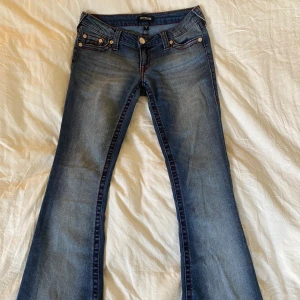 True Religion bootcut jeans blå - Snygga blå jeans från True Religion med bootcut passform och låg midja. Jeansen har klassiska röda kontrastsömmar, fem fickor och tydliga broderade bakfickor. Tillverkade i jeansmaterial med en cool tvättad look. Storlek 26.