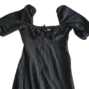 Svart maxi omlottklänning med puffärm - [KNAPPT ANVÄND] Supersnygg svart klänning från H&M med puffiga korta ärmar och omlottknytning. Klänningen har en lätt strukturerad yta, söt knytning vid bysten och dragkedja i sidan. Perfekt för dig som gillar trendiga detaljer och en feminin siluett.