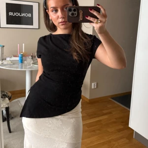 Svart figurnära topp med kort ärm - En stilren svart topp med korta ärmar och rund hals. Modellen är figurnära och har en lätt asymmetrisk nederkant som ger en cool twist. Perfekt att matcha med kjol eller jeans för en clean look.