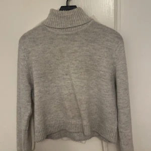 Grå stickad polotröja - Mysig grå stickad polotröja med ribbad hög krage och långa ärmar. Tröjan har en klassisk, enkel design och är perfekt för kyliga dagar. Den är mjuk och har ribbade muddar vid ärmslut och nederkant för extra komfort.