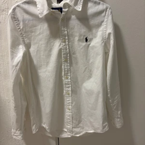 Vit skjorta från Ralph Lauren -  skjorta från Ralph Lauren med broderad logga på bröstet. Skjortan har lång ärm, knappar framtill och är tillverkad i mjuk bomull. Perfekt för en clean och stilren look. Sitter som s men är 16år