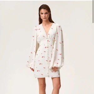 Adoore klänning - Superfin vit kortklänning med små röda blommor, djup v-ringning och långa puffärmar. Klänningen har markerad midja och knappar framtill, vilket ger en romantisk och trendig look. Perfekt för sommarens alla tillfällen! Använd en gång, 