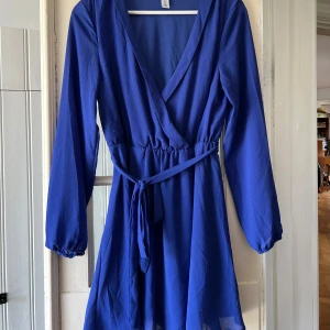 Blå omlottklänning från NLY Trend - Snygg blå omlottklänning från NLY Trend i storlek 38. Klänningen har långa ärmar, v-ringning och knytband i midjan för en smickrande passform. Perfekt för dig som vill ha en stilren och trendig look.