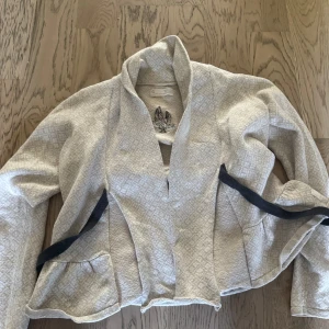 Beige cardigan jacka Odd Molly - Superfin beige cardigan jacka från Odd Molly med diskret rutigt mönster och broderad logga med fjädrar på ryggen. Jackan har lång ärm, öppen front och coola svarta banddetaljer vid fickorna. Perfekt för dig som gillar unika detaljer och mjuka material.