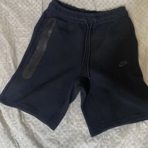 Svarta Nike shorts med snörning M - Svarta shorts från Nike med snygg minimalistisk design. De har snörning i midjan, dragkedjeficka på sidan och Nike-logga framtill. Perfekta för träning eller chill, tillverkade i mjukt och bekvämt material som känns skönt mot huden.