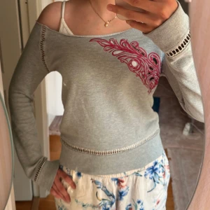 Grå offshoulder tröja med broderi - Grå offshoulder tröja med lång ärm och snygga håldetaljer vid ärmslut och midja. På bröstet finns ett broderat mönster i rosa som ger en unik touch. Tröjan har en relaxed passform och är perfekt för dig som gillar en chill men ändå stilren look.
