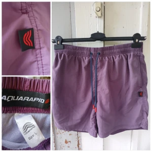 Lila badshorts från Aquarapid, strl L - Säljer ett par lila badshorts från Aquarapid i storlek L. Shortsen har elastisk midja med blå snörning och röda detaljer, innerbyxa i mesh och en bakficka. Perfekta för stranden eller poolen!