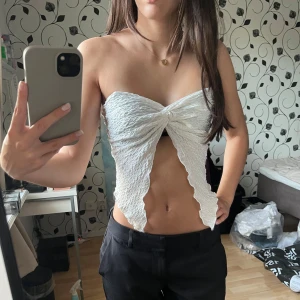 Vit bandeau tubtopp med twist - Säljer en vit bandeau tubtopp med twist framtill och vågiga kanter. Toppen är ärmlös och har en snygg struktur i tyget som ger en cool look. Perfekt för varma dagar, sommaren och utomlands. 