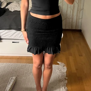 Svart tight kjol med volangkant - Supersnygg svart kortkjol med tight passform och volangdetalj längst ner. Kjolen är smockad och stretchig vilket gör den bekväm och sitter snyggt på kroppen. 