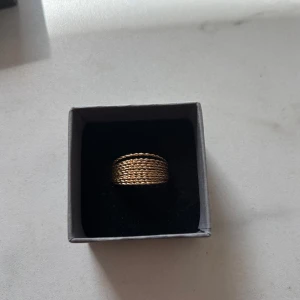 Flätad guldring - Säljer en snygg guldfärgad ring med flätad design. Man kan enkel vika flätningarna åt sidan och det har den ena gjort därför är den inte intill de andra. Men den är inte sönder. Vet inte om den rostar eller om man blir grön av den då jag inte hade den på mig länge! OBS! Asken medföljer ej! Läs gärna bio🥰