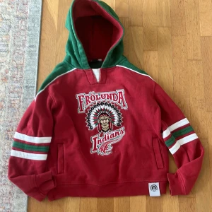 Frölunda Indians hoodie - Röd hoodie från Frölunda Indians med gröna och vita detaljer på ärmar och huva. Stor broderad logga framtill och sportig look. Perfekt för dig som älskar hockey och vill sticka ut med färg. Mjuk insida och klassisk känguruficka. Går ej att hitta längre