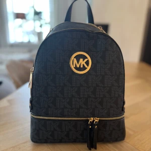 Svart ryggsäck från Michael Kors - Oanvänd, snygg svart ryggsäck från Michael Kors med guldiga detaljer och MK-logga framtill. Väskan har justerbara axelremmar, flera dragkedjefack och är täckt av diskret MK-mönster. Perfekt för dig som vill ha en stilren och trendig accessoar. Inköpspriset var 1000kr