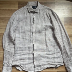 Beige linneskjorta från Riley - Snygg beige skjorta från Riley i 100% linne. Skjortan har klassisk krage, långa ärmar och knäppning framtill. Perfekt för en chill och stilren look under varmare dagar.