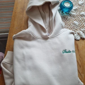 Beige hoodie Studio 1971 - Snygg beige hoodie från Studio 1971 med broderad grön logga på bröstet. Klassisk huva och ribbade muddar. Perfekt för en chill och avslappnad stil.
