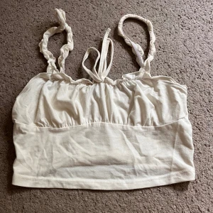 Vit croppad linnetopp med rynkade band - Supersöt vit croppad linnetopp med rynkade axelband och knytning framtill. Perfekt för varma dagar och enkel att matcha med jeans eller kjol. Mjuk och skön känsla tack vare bomullsmaterialet.