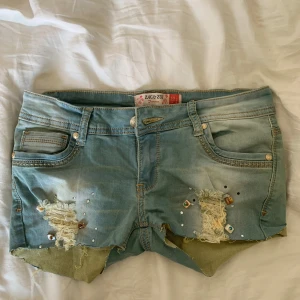 Jeansshorts med slitningar Zac & Zoe - Snygga ljusblå jeansshorts från Zac & Zoe med slitna detaljer, råa kanter och dekorativa nitar och pärlor framtill. Klassisk femficksmodell med låg midja och coola slitningar för en edgy look. Perfekta för sommaren! Det står storlek L men jag har s36 och de passar utmärkt