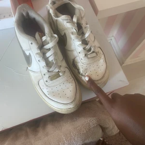 Nike Air Force 1 vita med gulddetaljer - Nike Air Force 1 sneakers i vitt skinn med metallic guld Swoosh och detaljer. Klassisk låg modell med perforerad tå, snörning och platt sula. Perfekta för dig som gillar retrovibe och streetstyle. Snygga att styla med jeans eller shorts, har en hål på framsidan. Ny pris 2500kr