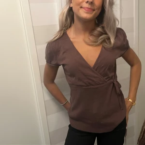 Brun omlottopp med knytband - Säljer en brun omlottopp med korta ärmar och v-ringning. Toppen har knytband i sidan och ribbad nederkant som ger en snygg passform. Perfekt till jeans eller kjol för en chill och stilren look.