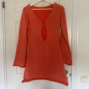 Glittrig orange stickad kortklänning - Snygg orange stickad kortklänning med glittriga trådar och lång ärm. Klänningen har v-ringning och en cool cut-out framtill med metallring som detalj. Perfekt för dig som vill sticka ut med färg och glitter.🧡Klänningen är aldrig använd i nyskick & fraktas 1-2 dagar ‼️